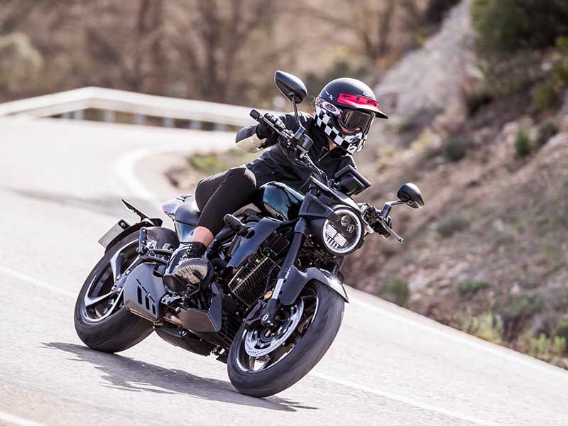 La foto muestra a una persona en una moto de carretera. La persona está inclinada hacia adelante y parece estar en movimiento, posiblemente acelerando. La imagen captura a un motorista montando una motocicleta negra y moderna. El motorista lleva un casco rojo y negro, y ropa oscura. La motocicleta está en movimiento, como se evidencia por la postura inclinada del motorista. Se encuentran en una carretera asfaltada que se curva suavemente hacia la izquierda. Al fondo, hay vegetación baja y árboles dispersos junto a la carretera, indicando un entorno rural o montañoso. El cielo es claro pero no es visible debido al ángulo de la cámara; sin embargo, la iluminación indica que es un día soleado.