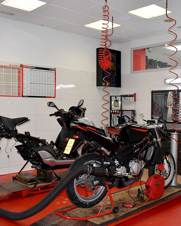 La foto muestra una motocicleta desmontada en un taller mecánico limpio y organizado. La imagen presenta un taller mecánico con paredes blancas y piso rojo. Una motocicleta negra está parcialmente desmontada y colocada sobre una plataforma elevadora. Hay herramientas y equipos organizados, incluyendo una manguera de aire comprimido que cuelga del techo. Dos tableros con horarios o listas están montados en la pared, aunque el texto no es legible. Un extractor de humos negro está conectado a la motocicleta para ventilar los gases. Hay un armario de herramientas gris a la derecha con cajones etiquetados, indicando un ambiente organizado.
