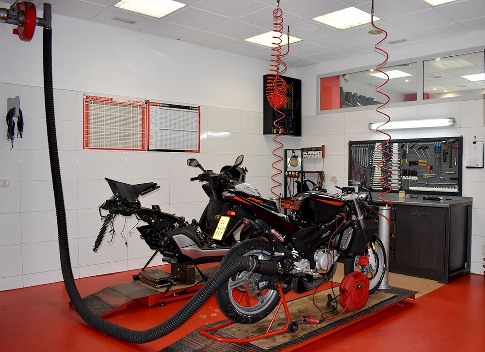 La foto muestra una motocicleta desmontada en un taller mecánico limpio y organizado. La imagen presenta un taller mecánico con paredes blancas y piso rojo. Una motocicleta negra está parcialmente desmontada y colocada sobre una plataforma elevadora. Hay herramientas y equipos organizados, incluyendo una manguera de aire comprimido que cuelga del techo. Dos tableros con horarios o listas están montados en la pared, aunque el texto no es legible. Un extractor de humos negro está conectado a la motocicleta para ventilar los gases. Hay un armario de herramientas gris a la derecha con cajones etiquetados, indicando un ambiente organizado.